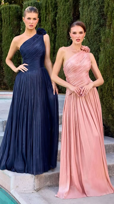 Two women in pleated, floor-length evening gowns by a pool: navy blue one-shoulder dress with a flower detail and blush pink one-shoulder dress with a matching flower.