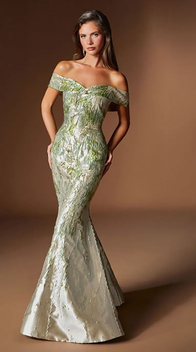 Full-length woman in an off-shoulder, emerald-green mermaid gown with a floral lace pattern posing with hands on hips against a brown backdrop.