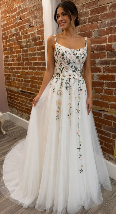 Woman wearing a white floral-embroidered wedding dress, standing against a brick wall indoors.