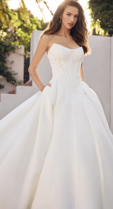 Bride in a strapless white ball gown with an embroidered bodice, standing outdoors with hands lightly in the dress pockets.