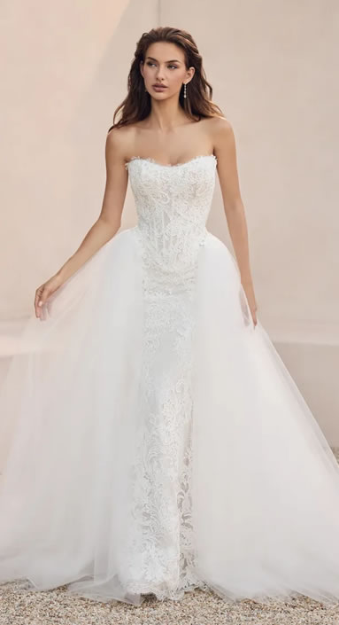 Bride modeling a strapless white lace gown with a fitted bodice and a voluminous tulle overskirt, standing outdoors on a light background.
