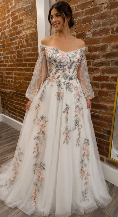 Bride in an off-the-shoulder wedding gown with sheer long sleeves and pastel floral embroidery, standing against a brick wall in a fitting room.